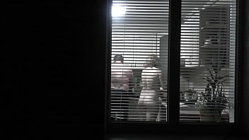 Peeping. Voyeur. Neighbor pervert voyeur in evening on street looks out window as naked sexy neighbor in kitchen prepares dinner for her husband. Naked in public. Naked at home. Family.  Outdoor